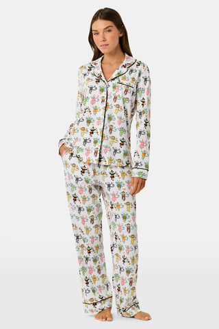 A smiling child in Roller Rabbit’s Kids Boo Crew Pajamas, crafted from soft Pima cotton, sits with arms raised on outdoor steps among women in coordinating patterned pajamas, all sharing a cheerful and relaxed moment together.