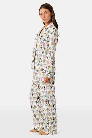 A woman stands barefoot against a plain white background, wearing the Roller Rabbit Boo Crew Long Sleeve Polo Pajamas, which feature a colorful cartoon bee and flower pattern on white Pima cotton with long sleeves and button-up styling.