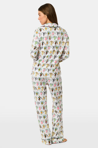 A woman stands barefoot against a plain white background, wearing the Roller Rabbit Boo Crew Long Sleeve Polo Pajamas, which feature a colorful cartoon bee and flower pattern on white Pima cotton with long sleeves and button-up styling.