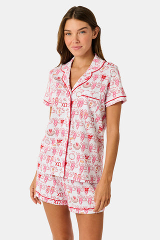 A young woman with red hair sits on a bench outside, holding a phone and earphones while looking to the side. She is wearing the Roller Rabbit Chi Omega Monkey Polo Pajamas, which feature a front button placket and an elastic waistband in a pink hue. A light jacket is draped over the bench beside her.