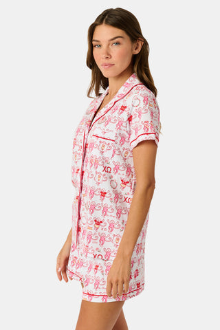 A young woman with red hair sits on a bench outside, holding a phone and earphones while looking to the side. She is wearing the Roller Rabbit Chi Omega Monkey Polo Pajamas, which feature a front button placket and an elastic waistband in a pink hue. A light jacket is draped over the bench beside her.