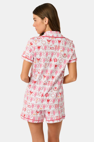 A young woman with red hair sits on a bench outside, holding a phone and earphones while looking to the side. She is wearing the Roller Rabbit Chi Omega Monkey Polo Pajamas, which feature a front button placket and an elastic waistband in a pink hue. A light jacket is draped over the bench beside her.