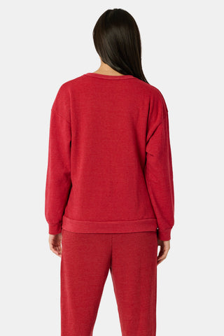 A smiling woman sits on a wooden bench wearing a red Deck The Halls Andie Sweatshirt featuring an illustrated monkey in string lights and patterned Bea Sweatpants.