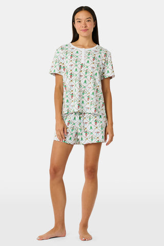 A woman wears a SERFLEX Deck The Halls Bailey Pajamas set featuring a short-sleeved top and shorts with a white background and a repeating Christmas-themed pattern of elves, snowmen, and evergreen trees.