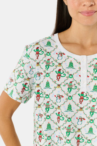 A woman wears a SERFLEX Deck The Halls Bailey Pajamas set featuring a short-sleeved top and shorts with a white background and a repeating Christmas-themed pattern of elves, snowmen, and evergreen trees.