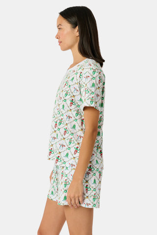 A woman wears a SERFLEX Deck The Halls Bailey Pajamas set featuring a short-sleeved top and shorts with a white background and a repeating Christmas-themed pattern of elves, snowmen, and evergreen trees.