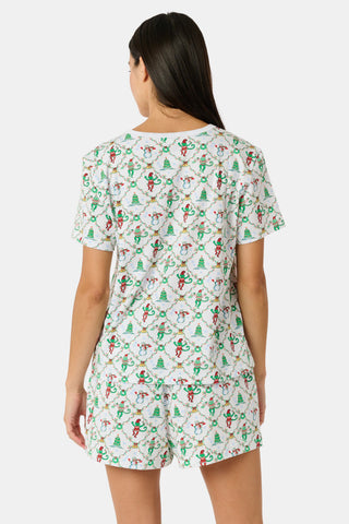 A woman wears a SERFLEX Deck The Halls Bailey Pajamas set featuring a short-sleeved top and shorts with a white background and a repeating Christmas-themed pattern of elves, snowmen, and evergreen trees.