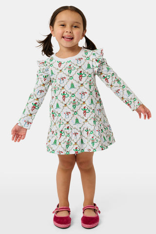 A young girl wears a Kids Deck The Halls Isa Dress, a long-sleeved Pima Cotton holiday dress featuring a white background with a festive monkey print of snowmen and Christmas trees.