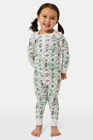 A child wearing SERFLEX Kids Deck The Halls Pajamas, featuring a repeating holiday pattern of snowmen, Christmas trees, and elves connected by garland on long-sleeved Pima cotton.