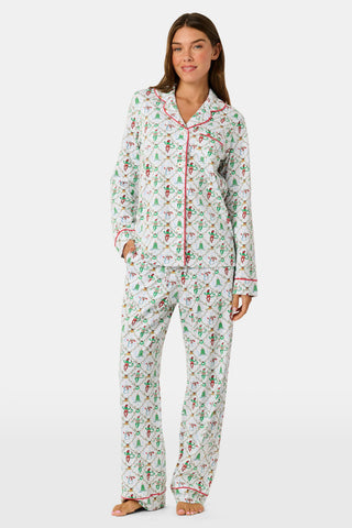 A woman wearing SERFLEX Deck The Halls Long Sleeve Polo Pajamas, a two-piece Pima cotton set featuring a repeating Christmas print of snowmen, elves, and trees on a white background with red piping.