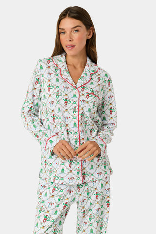 A woman wearing SERFLEX Deck The Halls Long Sleeve Polo Pajamas, a two-piece Pima cotton set featuring a repeating Christmas print of snowmen, elves, and trees on a white background with red piping.