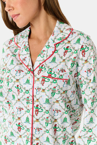 A woman wearing SERFLEX Deck The Halls Long Sleeve Polo Pajamas, a two-piece Pima cotton set featuring a repeating Christmas print of snowmen, elves, and trees on a white background with red piping.