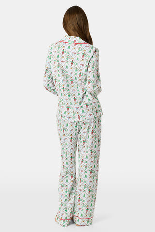 A woman wearing SERFLEX Deck The Halls Long Sleeve Polo Pajamas, a two-piece Pima cotton set featuring a repeating Christmas print of snowmen, elves, and trees on a white background with red piping.