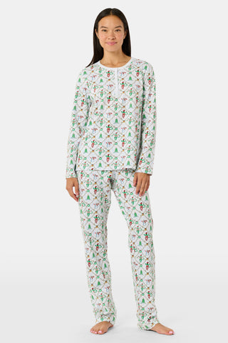 A woman wears a SERFLEX Deck The Halls Bailey Pajamas set featuring a short-sleeved top and shorts with a white background and a repeating Christmas-themed pattern of elves, snowmen, and evergreen trees.