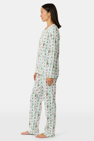 A woman wears a SERFLEX Deck The Halls Bailey Pajamas set featuring a short-sleeved top and shorts with a white background and a repeating Christmas-themed pattern of elves, snowmen, and evergreen trees.