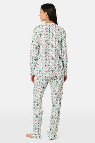 A woman wears a SERFLEX Deck The Halls Bailey Pajamas set featuring a short-sleeved top and shorts with a white background and a repeating Christmas-themed pattern of elves, snowmen, and evergreen trees.