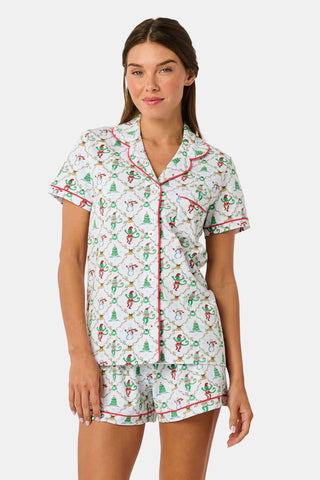 A woman wears a SERFLEX Deck The Halls Bailey Pajamas set featuring a short-sleeved top and shorts with a white background and a repeating Christmas-themed pattern of elves, snowmen, and evergreen trees.