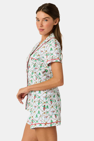 A woman wears a SERFLEX Deck The Halls Bailey Pajamas set featuring a short-sleeved top and shorts with a white background and a repeating Christmas-themed pattern of elves, snowmen, and evergreen trees.
