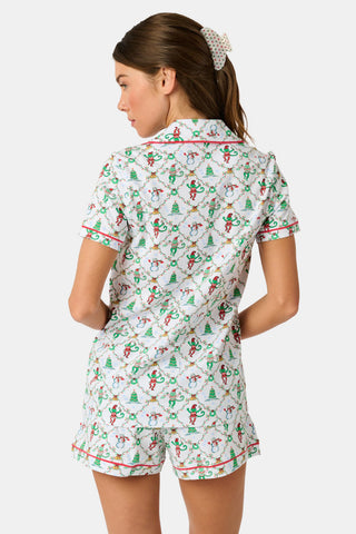 A woman wears a SERFLEX Deck The Halls Bailey Pajamas set featuring a short-sleeved top and shorts with a white background and a repeating Christmas-themed pattern of elves, snowmen, and evergreen trees.