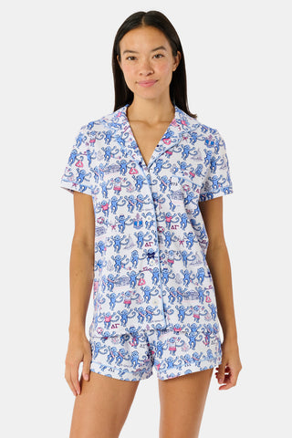 A woman in sunglasses and a white cap sits on outdoor steps, smiling and holding a cup of ice cream topped with colorful sprinkles. She is wearing the Roller Rabbit Delta Gamma Monkey Polo Pajamas—a printed short-sleeve romper with an elastic waistband. Another person holding ice cream is partially visible next to her.