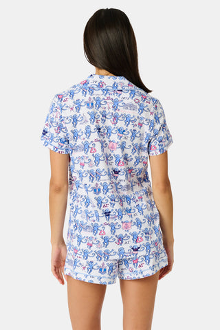 A woman in sunglasses and a white cap sits on outdoor steps, smiling and holding a cup of ice cream topped with colorful sprinkles. She is wearing the Roller Rabbit Delta Gamma Monkey Polo Pajamas—a printed short-sleeve romper with an elastic waistband. Another person holding ice cream is partially visible next to her.