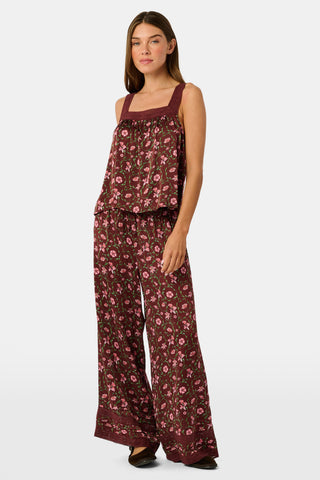 A woman wears a burgundy Greta Floral Ditsy Rowan Set camisole and wide-leg trousers, standing in front of a rustic wooden fence decorated with Christmas stockings.