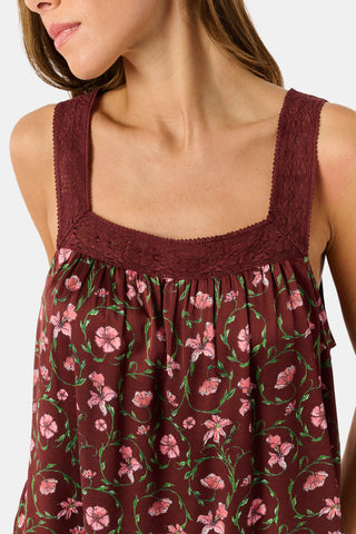 A woman wears a burgundy Greta Floral Ditsy Rowan Set camisole and wide-leg trousers, standing in front of a rustic wooden fence decorated with Christmas stockings.