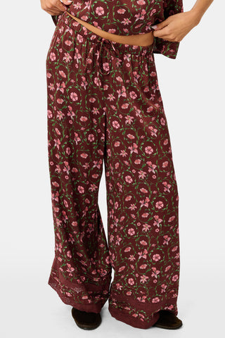 A woman wears a burgundy Greta Floral Ditsy Rowan Set camisole and wide-leg trousers, standing in front of a rustic wooden fence decorated with Christmas stockings.