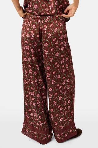 A woman wears a burgundy Greta Floral Ditsy Rowan Set camisole and wide-leg trousers, standing in front of a rustic wooden fence decorated with Christmas stockings.