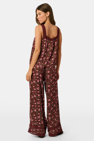 A woman wears a burgundy Greta Floral Ditsy Rowan Set camisole and wide-leg trousers, standing in front of a rustic wooden fence decorated with Christmas stockings.