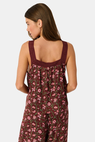 A woman wears a burgundy Greta Floral Ditsy Rowan Set camisole and wide-leg trousers, standing in front of a rustic wooden fence decorated with Christmas stockings.