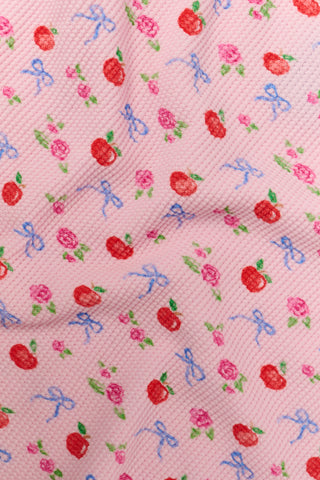 A young girl stands smiling, wearing the Roller Rabbit Kids Apple Bloom Elle Set—a pink girls’ loungewear outfit featuring an apple bloom print and a ruffled hem. She has long dark hair and is wearing light pink shoes, set against a plain white background.
