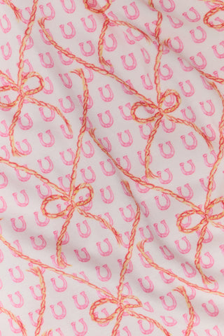 Wearing Roller Rabbit’s Lasso Bows Hayden Pajamas in pastel pink, a woman pairs the two-piece set with brown cowboy boots as she stands by a light-colored house door, smiling and resting one arm above her head while bending one leg with her foot on a step.