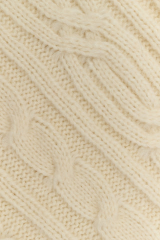 The Ruby Cardigan by REVOLUTION KNITS is a cream-colored, alpaca blend cable-knit cardigan with a crew neck and button front, worn over a red and white patterned skirt.