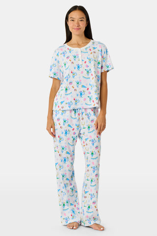 A woman with long blonde hair sits thoughtfully on an outdoor bench, surrounded by green foliage, wearing Roller Rabbit’s Game Day Evie Pajamas in a pastel print and holding a polka dot cup.
