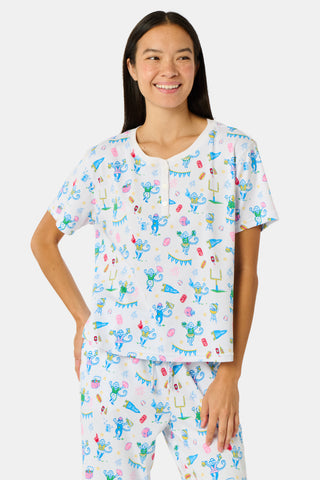 A woman with long blonde hair sits thoughtfully on an outdoor bench, surrounded by green foliage, wearing Roller Rabbit’s Game Day Evie Pajamas in a pastel print and holding a polka dot cup.
