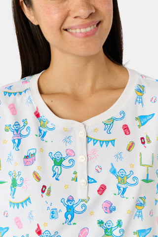 A woman with long blonde hair sits thoughtfully on an outdoor bench, surrounded by green foliage, wearing Roller Rabbit’s Game Day Evie Pajamas in a pastel print and holding a polka dot cup.