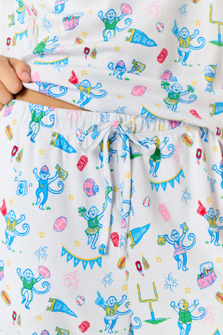 A woman with long blonde hair sits thoughtfully on an outdoor bench, surrounded by green foliage, wearing Roller Rabbit’s Game Day Evie Pajamas in a pastel print and holding a polka dot cup.