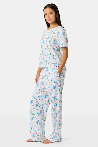 A woman with long blonde hair sits thoughtfully on an outdoor bench, surrounded by green foliage, wearing Roller Rabbit’s Game Day Evie Pajamas in a pastel print and holding a polka dot cup.