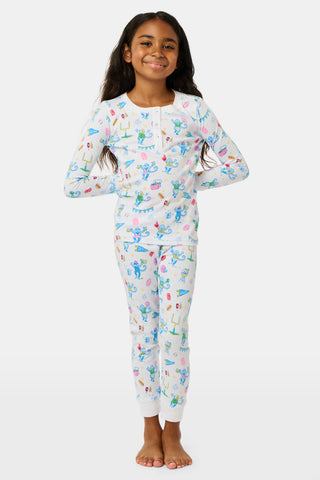 The Kids Game Day Pajamas by Roller Rabbit are a children’s long-sleeve pajama set crafted from soft Pima cotton, featuring a vibrant party-themed print with colorful dinosaurs, bunting, gifts, and party hats on a white background.