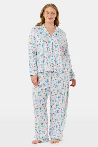 Roller Rabbit’s Game Day Long Sleeve Polo Pajamas feature a white button-up shirt and matching pants adorned with colorful, playful animal prints, festive bunting, and other cheerful designs.