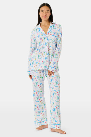 Roller Rabbit’s Game Day Long Sleeve Polo Pajamas feature a white button-up shirt and matching pants adorned with colorful, playful animal prints, festive bunting, and other cheerful designs.