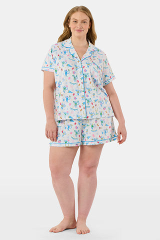 The Game Day Polo Pajamas by Roller Rabbit feature a short-sleeve button-up top and shorts, adorned with a lively pattern of colorful cartoon animals and party items on a white background.