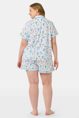 The Game Day Polo Pajamas by Roller Rabbit feature a short-sleeve button-up top and shorts, adorned with a lively pattern of colorful cartoon animals and party items on a white background.
