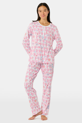 A woman wearing Roller Rabbit Gamma Phi Beta Monkey Pajamas—featuring a white and pink pattern with an elastic waistband—stands outdoors in front of greenery, holding a phone to her ear, a tumbler, and keys. A sweatshirt is tied around her waist, and she wears sunglasses.