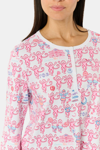 A woman wearing Roller Rabbit Gamma Phi Beta Monkey Pajamas—featuring a white and pink pattern with an elastic waistband—stands outdoors in front of greenery, holding a phone to her ear, a tumbler, and keys. A sweatshirt is tied around her waist, and she wears sunglasses.
