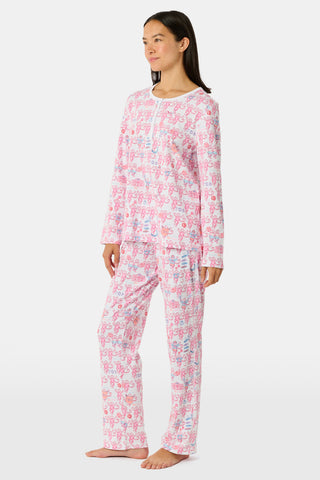 A woman wearing Roller Rabbit Gamma Phi Beta Monkey Pajamas—featuring a white and pink pattern with an elastic waistband—stands outdoors in front of greenery, holding a phone to her ear, a tumbler, and keys. A sweatshirt is tied around her waist, and she wears sunglasses.