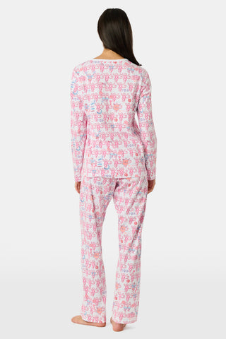 A woman wearing Roller Rabbit Gamma Phi Beta Monkey Pajamas—featuring a white and pink pattern with an elastic waistband—stands outdoors in front of greenery, holding a phone to her ear, a tumbler, and keys. A sweatshirt is tied around her waist, and she wears sunglasses.
