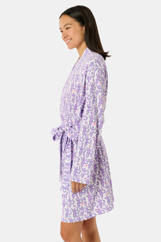 A woman smiles while wearing the TEXPIMA Get Ready With Me Blake Robe, featuring a knee-length cut and a purple and pink paisley pattern, tied at the waist, against a plain white background.