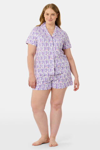 A woman is wearing the TEXPIMA Get Ready With Me Polo Pajamas — a matching short-sleeve button-up top and shorts set featuring a purple and pink patterned print — while standing against a plain light background, looking at the camera with a gentle smile.